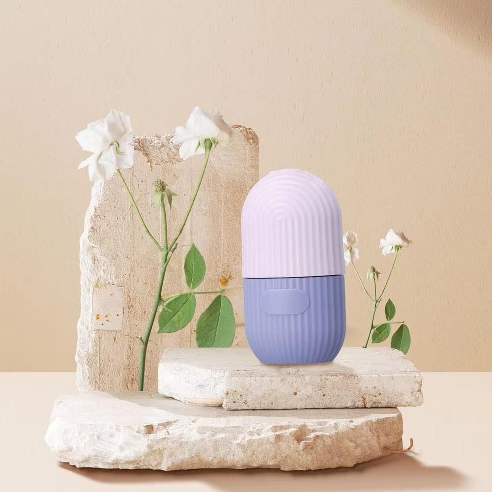 The Face Ice Lattice – Ice Roller Face Massager, in pastel purple and light pink, stands on stone slabs amid delicate white flowers and green leaves against a beige background.